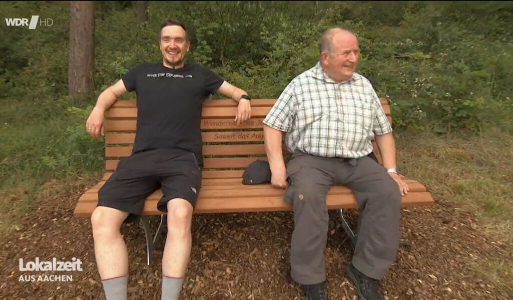 Die EifelSpur „So weit das Auge reicht“ wurde 2024 als Wanderweg des Jahres ausgezeichnet. Als Preis gab es eine Bank. David Rosenbaum (l.) und Horst Müller nahmen für einen WDR-Bericht dort Platz. Screenshot: Ronald Larmann/pp/Agentur ProfiPress