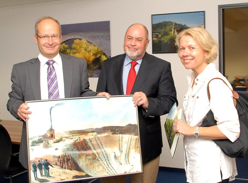 Birgit und Klaus Vater zu Besuch bei Bürgermeister Dr. Hans-Peter Schick im Mechernicher Rathaus. Sie halten ein berühmtes Bild, das Arbeitsmethoden im Tagebau „Virginia“ festhält, die an Sklaven auf einer Galeere erinnern. Die Berliner Gäste auf dem Gemälde gaben dem Bleiberg den Namen „Spandau“, der an die preußische Strafanstalt nahe Berlin erinnerte. Archivfoto: Manfred Lang/pp/Agentur ProfiPress