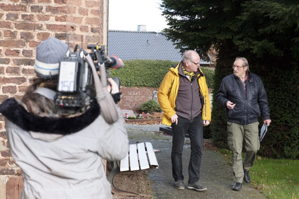 Vor laufender Fernsehkamera berichteten Kirchenvorstand Hans-Joachim Emonds (v.r.) und Pfarrer Erik Pühringer über den baufälligen Glockenturm und Dachstuhl der Holzheimer Pfarrkirche St. Lambertus. Beiden ist mit zu verdanken, dass die jahrelange Sanierung letztendlich geglückt ist, ohne dass das Unternehmen ein Fass ohne Boden wurde. Foto: Ron Larmann/pp/Agentur ProfiPress