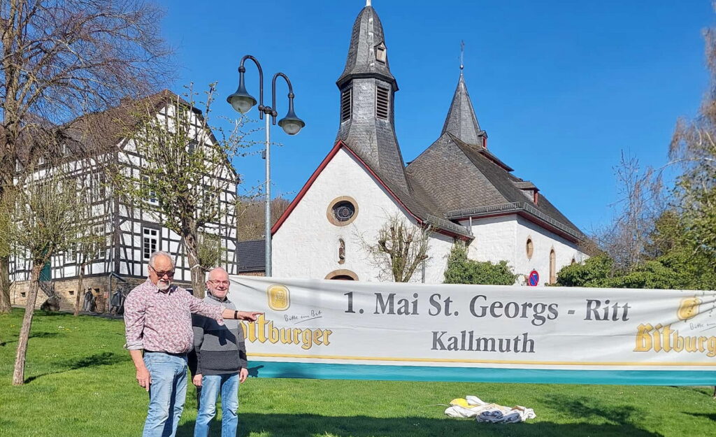Frühzeitig hängen Robert Ohlerth und Gerhard Mayr-Reineke (v.l.) die Transparente für den 72. Sankt-Georgsritt am Freitag, 1. Mai, um 11.15 Uhr in den Dörfern rings um den Mechernicher Bleiberg auf, hier vor der Pfarrkirche St. Wendelinus in Eiserfey. Foto: Hubert Hufschmidt/pp/Agentur ProfiPress
