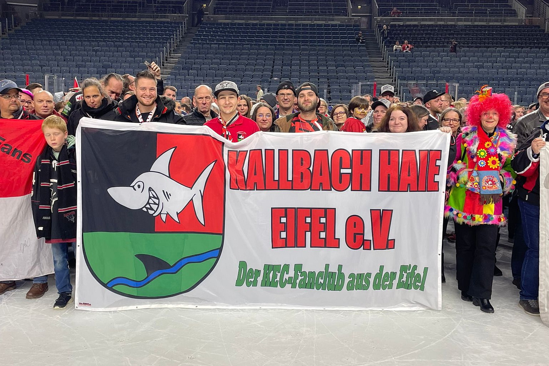 Die Eifel auf internationalem Parkett: Mit ihrem Vereinsbanner präsentieren sich die „Kallbach Haie Eifel e.V.“ bei einer Fanveranstaltung. Das von Tochter Anne May gestaltete Logo verbindet die Farben der Kölner Haie mit der grünen Landschaft der Eifel und dem namensgebenden Kallbach, aus dem eine Rückenflosse ragt. Foto: Privat/pp/Agentur ProfiPress