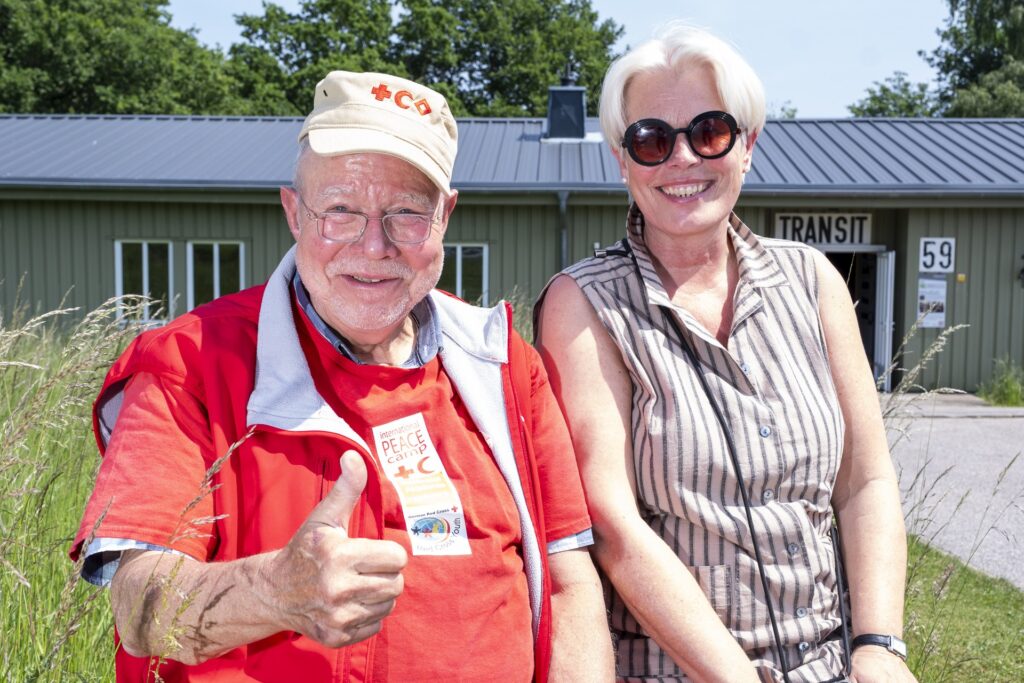 So werden ihn viele in Erinnerung behalten: fröhlich, positiv und mit einem verschmitzten Lächeln. Rolf Zimmermann im Juni 2025 mit Dr. Frauke Hartung von der befreundeten DRK-Schwesternschaft „Bonn“ e.V. vor „Transit 59“, einem der Gebäude in Vogelsang, die er über viele Jahre mit dem Geist und der Idee des Roten Kreuzes erfüllt hat. Und natürlich trägt er dabei ein Peace-Camp-T-Shirt. Foto: Ronald Larmann/pp/Agentur ProfiPress